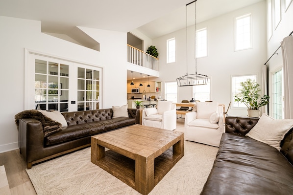 A spacious living room with a brown leather sofa, a wooden coffee table, and a white armchair. A kitchen is visible in the background, and there are large windows providing natural light. - Beautiful Luxury Home located at Island Pointe Marina!
Just 1 mile to downtown! (St. Joseph)