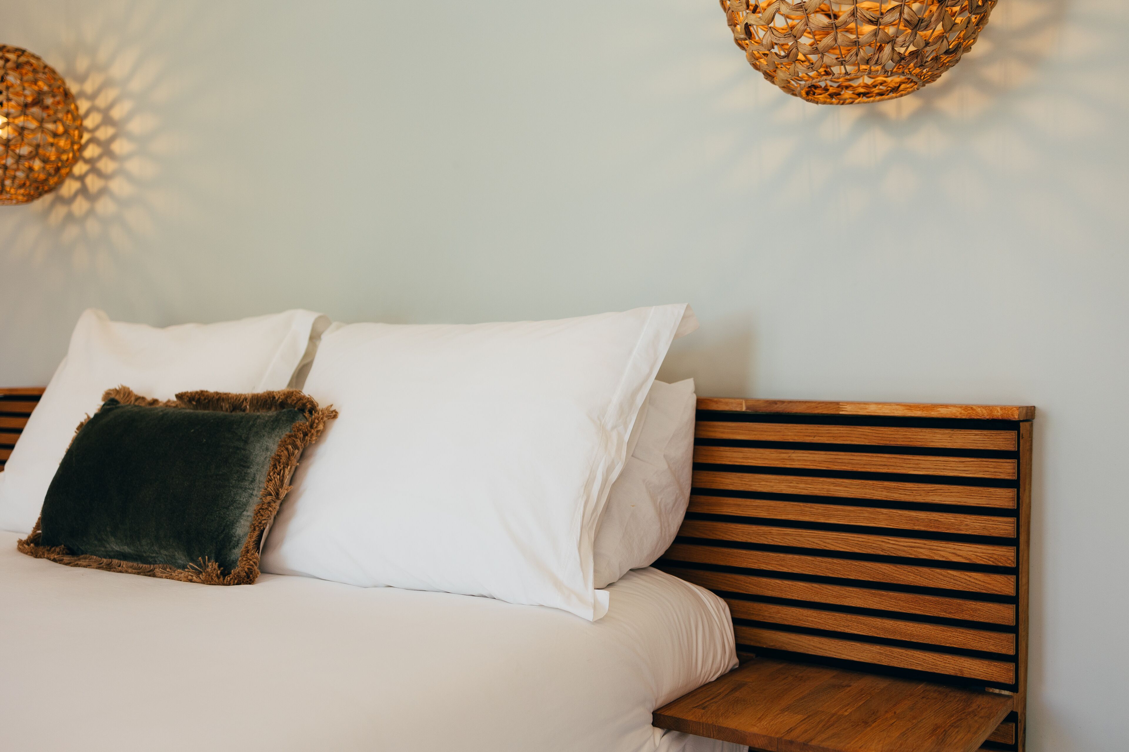 Una cama con sábanas blancas y un almohadón marrón oscuro, un cabecero de madera y una mesita de noche de madera.