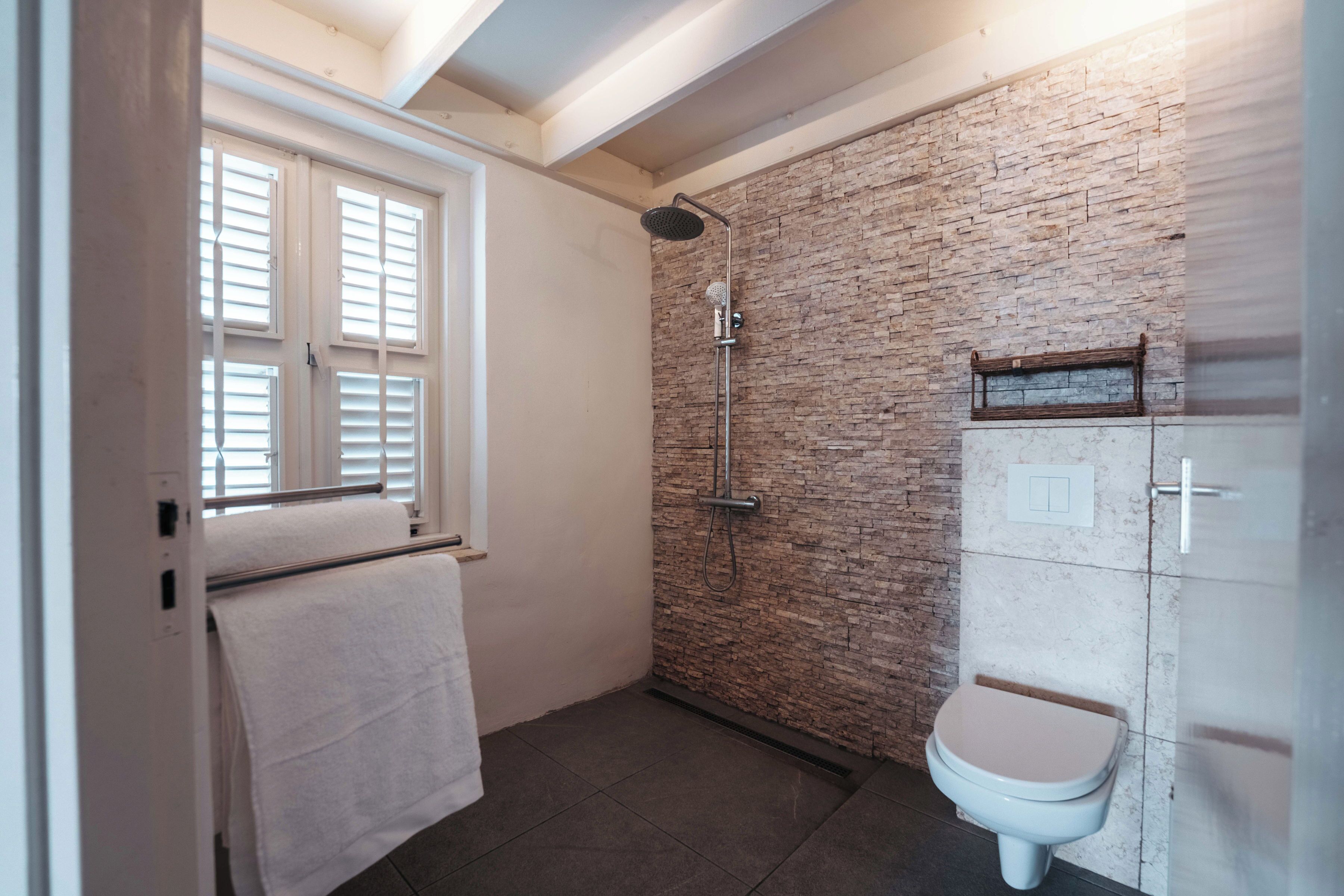 A bathroom with a shower, toilet, and a window with blinds.