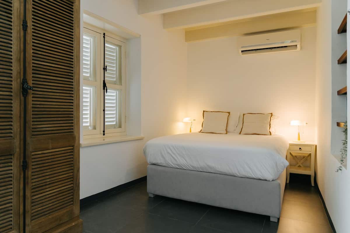 A bedroom with a bed, two bedside lamps, a window with shutters, and an air conditioning unit.