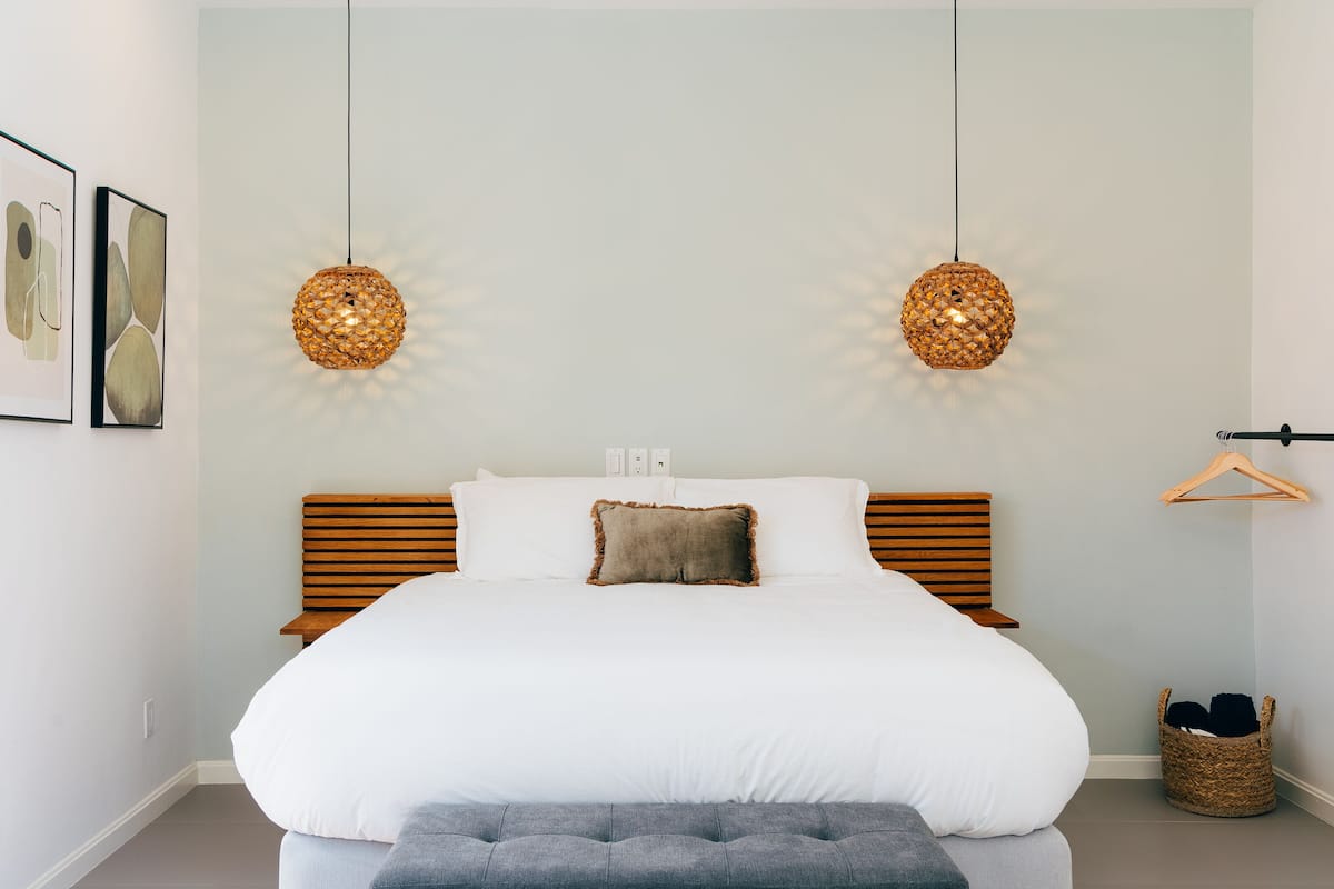 A bedroom with a large bed, two hanging lamps, a wooden headboard, a grey bench, and a basket on the floor.