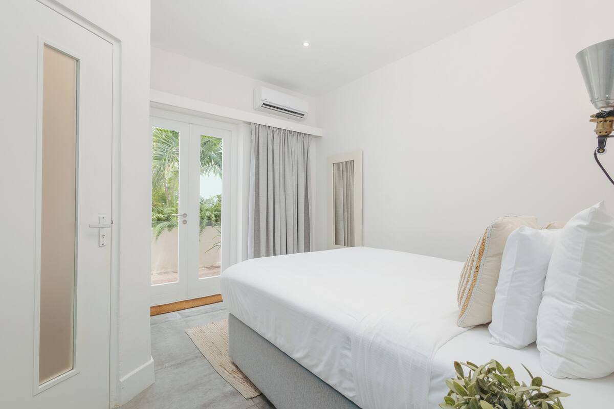 A bedroom with a bed, an air conditioning unit, a window with curtains, and a view of greenery outside.