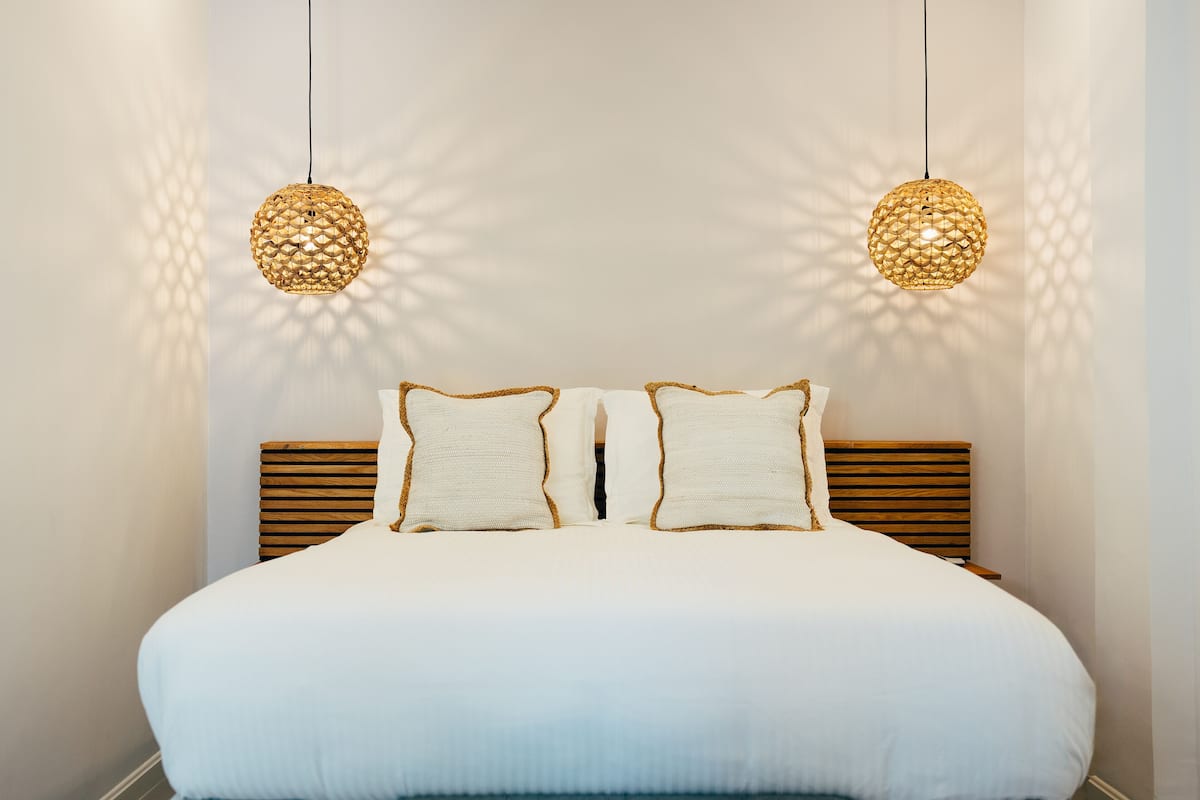 A neatly made bed with white bedding and two decorative pillows, a wooden headboard, and two hanging spherical light fixtures.