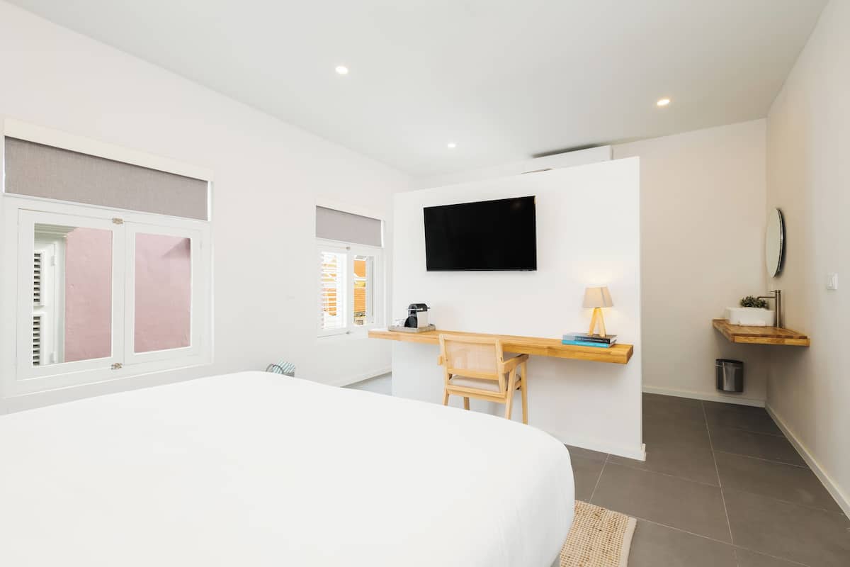 A modern hotel room with a large bed, a desk with a chair, a flat-screen TV mounted on the wall, and a small shelf with a lamp and a plant.