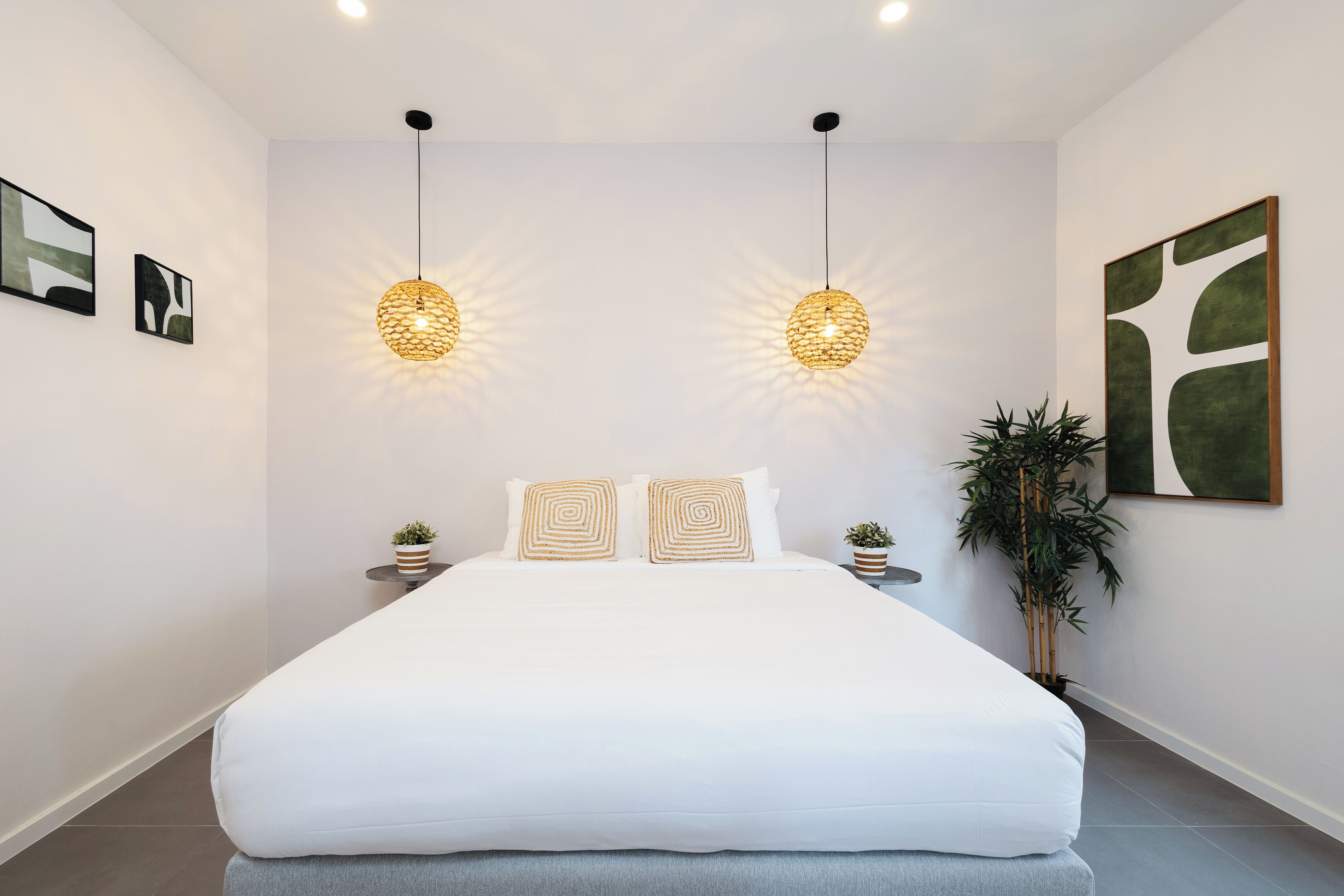 A modern bedroom with a large bed, two hanging pendant lights, a small potted plant, and a large abstract painting on the wall.
