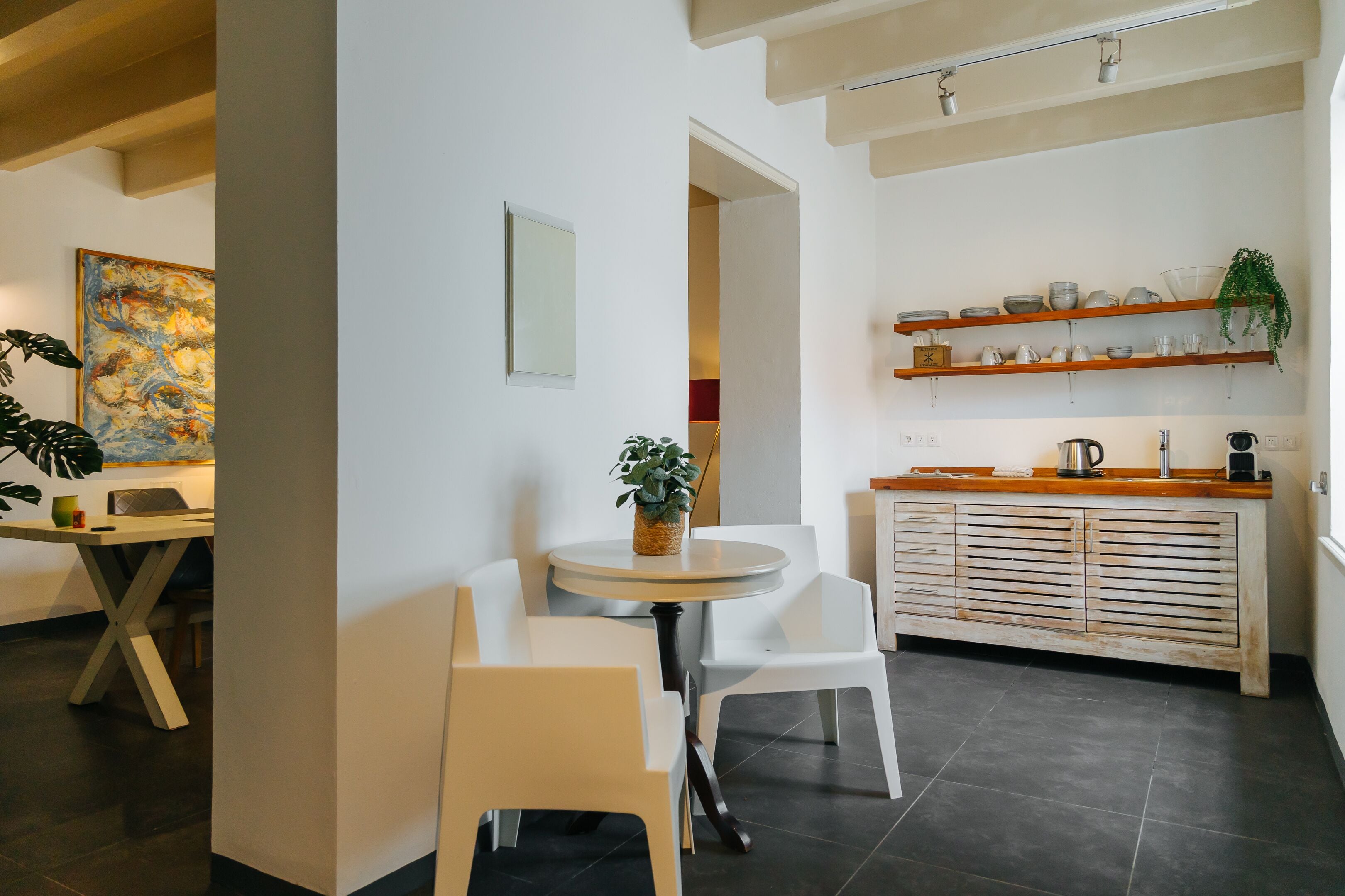 A modern kitchen with white cabinetry, a round dining table, and chairs. There are shelves with dishes and a potted plant.