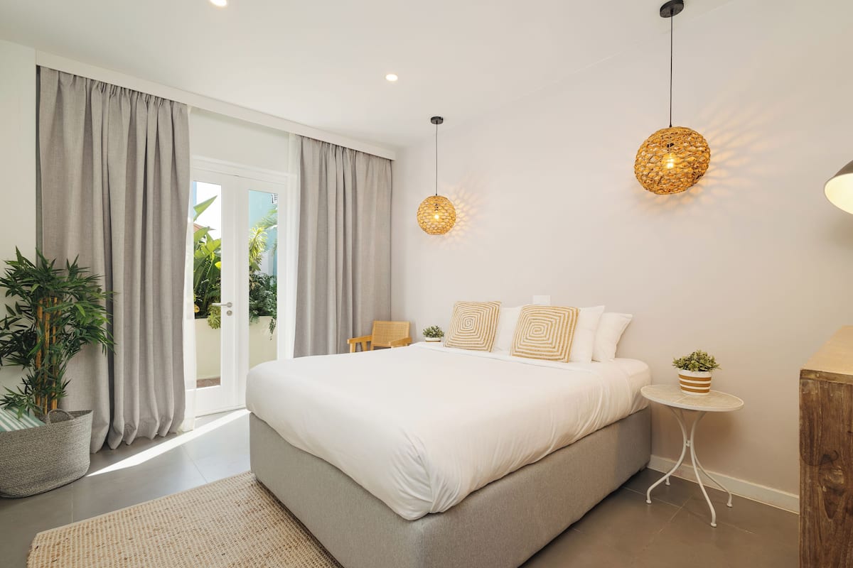 A bedroom with a large bed, a small round table, a potted plant, and a view of the outdoors through a sliding door.