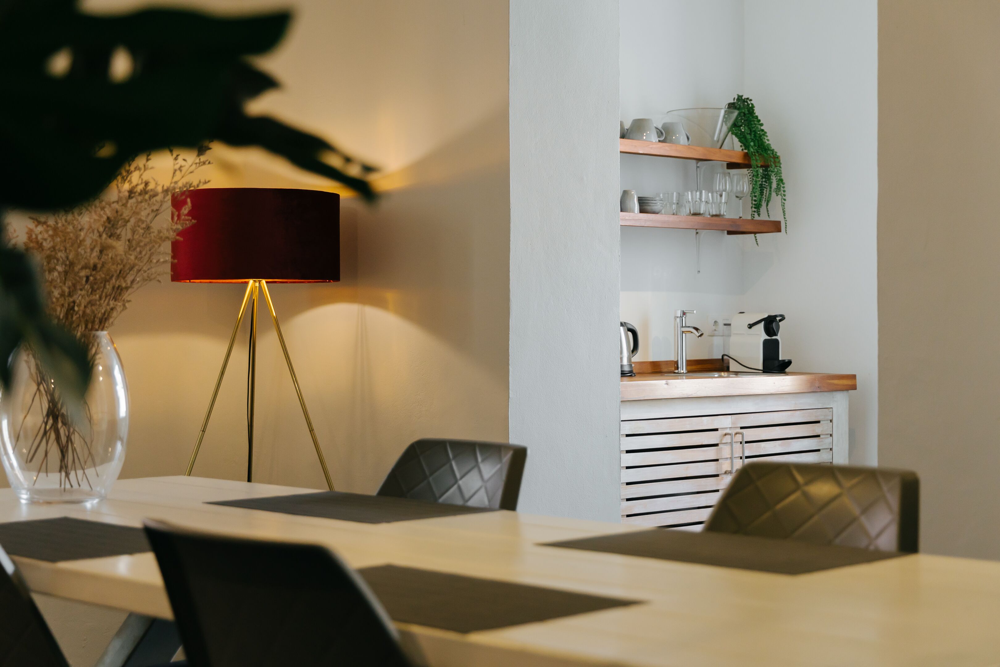 A modern dining area with a table, chairs, a red lamp, and a kitchen counter with shelves.