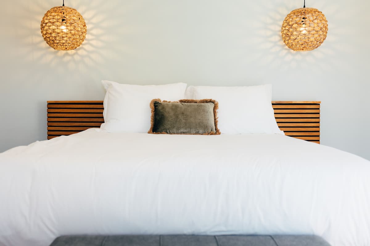 A neatly made bed with white bedding and two decorative pillows, a wooden headboard, and two hanging spherical light fixtures.