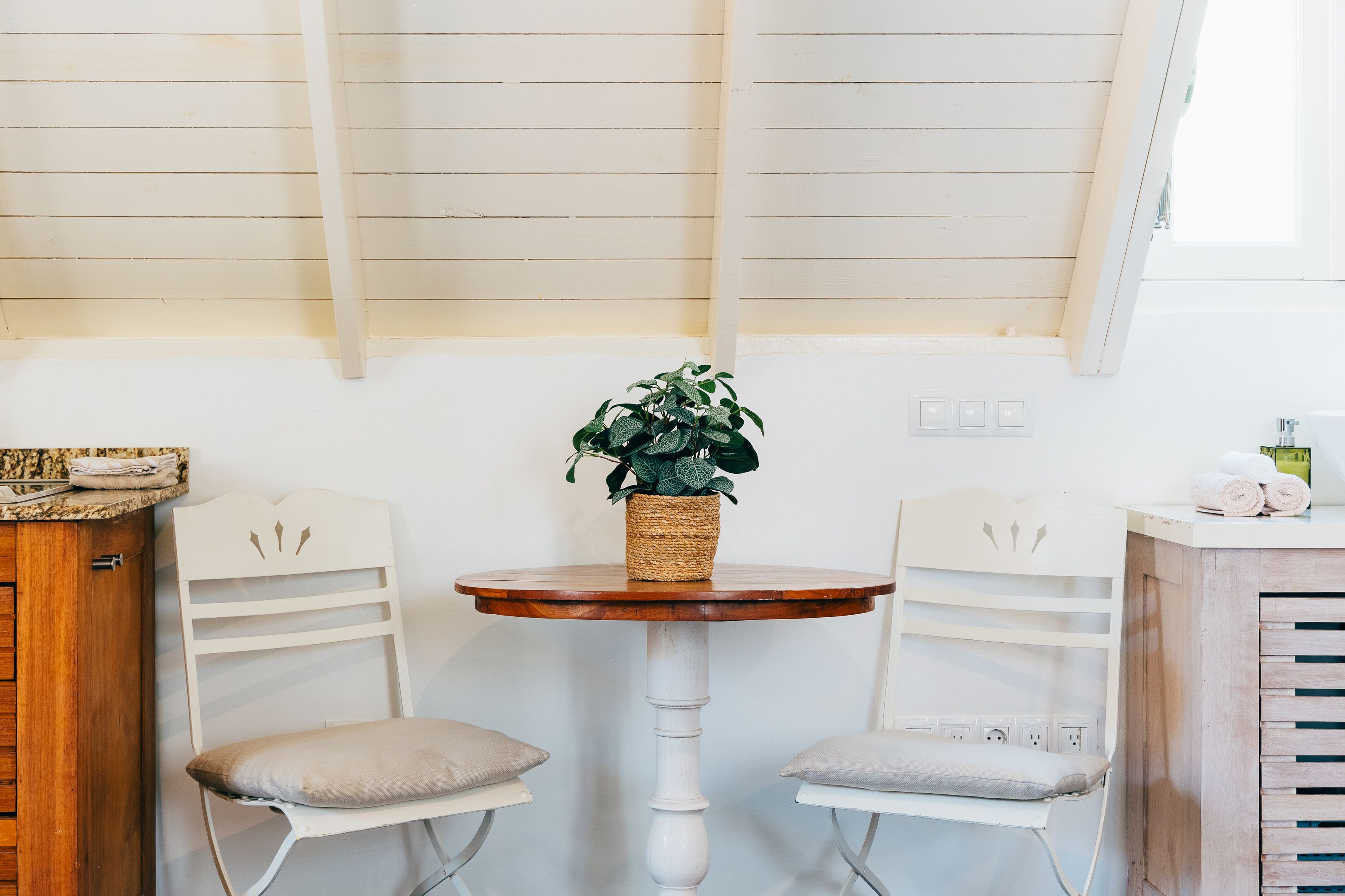Um espaço de refeições pequeno com uma mesa redonda de madeira, duas cadeiras brancas e um vaso com planta.
