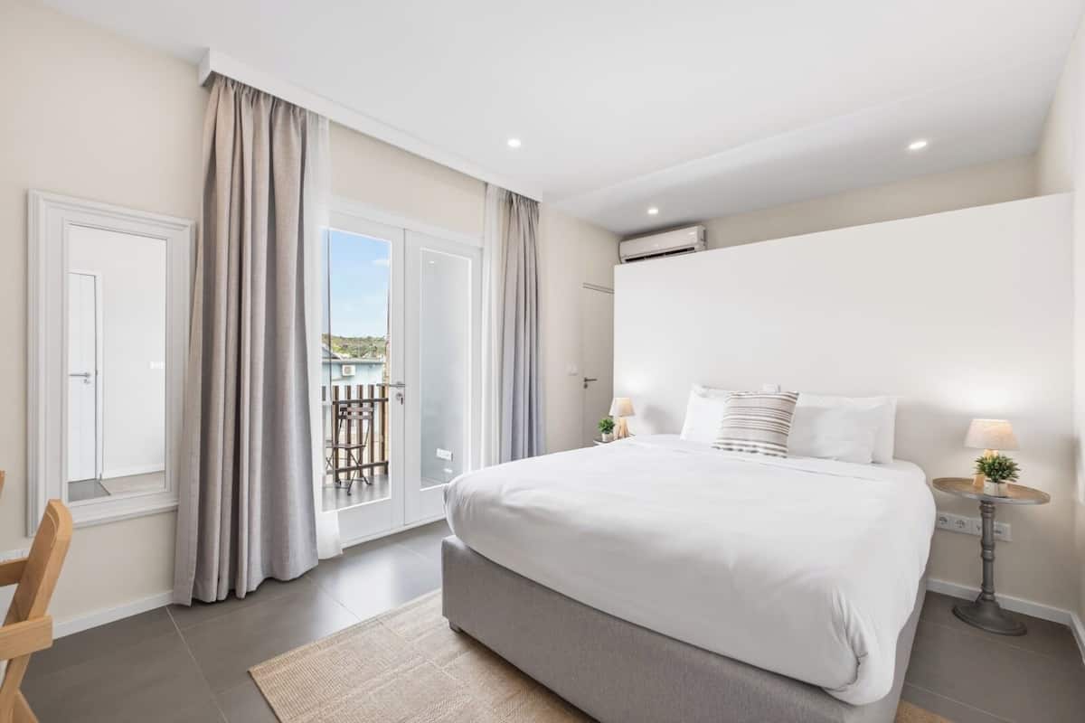 A bedroom with a large bed, a balcony with curtains, a window, and an air conditioning unit.