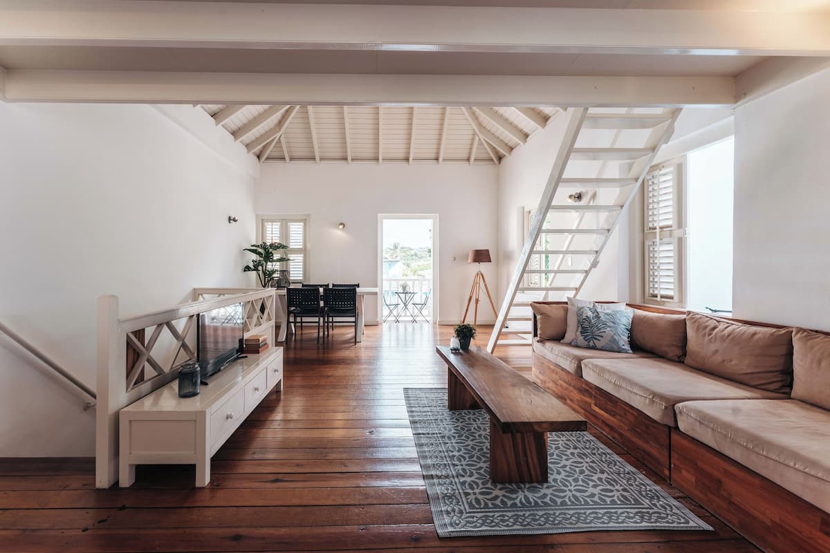 A spacious living area with a wooden floor, a sofa, a bench, and a TV on a white stand. There is a staircase leading to an upper level, and a door leading outside.