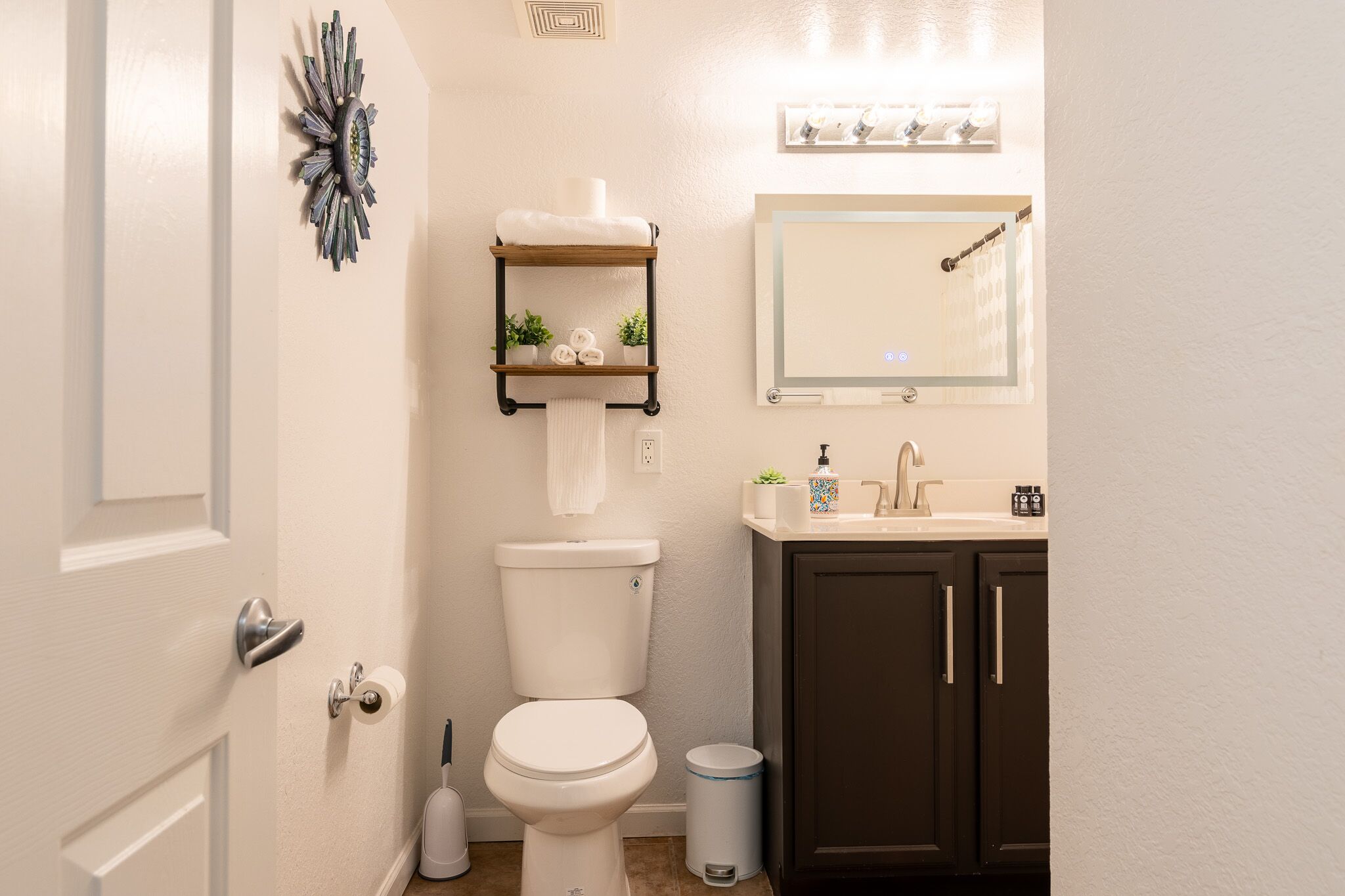 A bathroom with a toilet, sink, mirror, and a shelf with towels and plants.