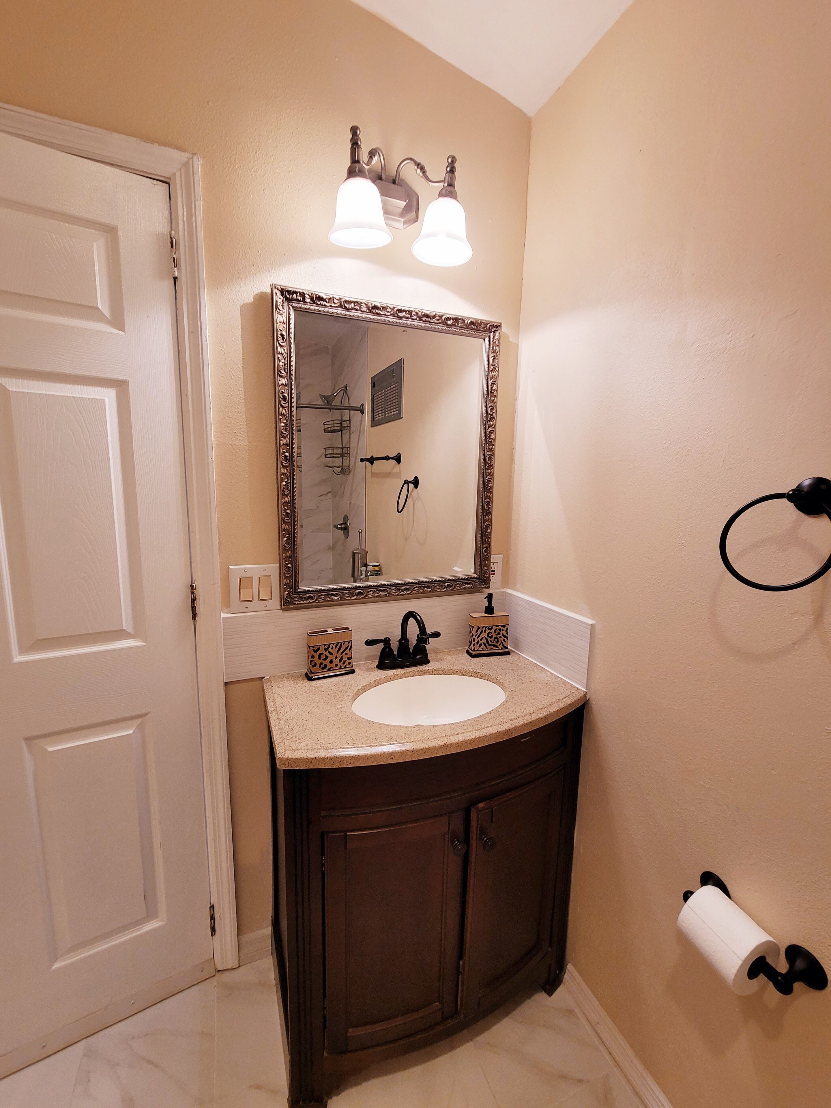 A bathroom with a white sink, dark wooden vanity, a large mirror with lights, a towel ring, and a toilet paper holder.