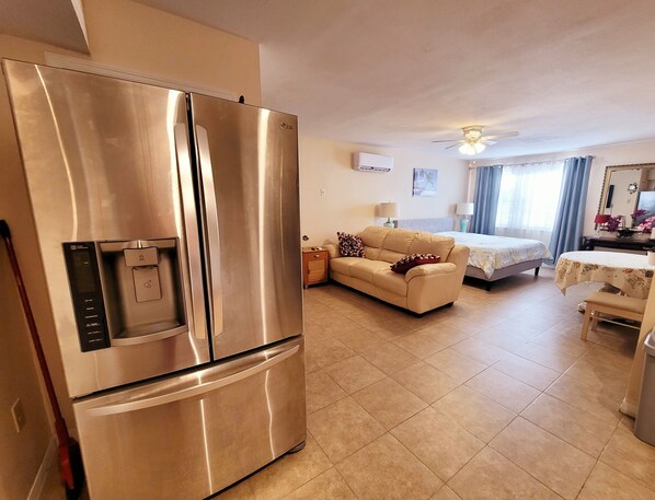 A modern kitchen with a stainless steel refrigerator, a dining area with a table and chairs, a sofa, and a bed in the background.