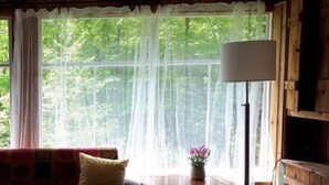 Une pièce chaleureuse avec un plafond en bois, un canapé agrémenté de coussins, une petite table avec une lampe, et une vue sur la verdure à l’extérieur.