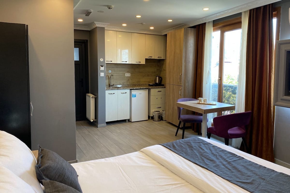 A compact hotel room with a bed, a small desk, a chair, and a kitchenette.