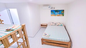 Una habitación pequeña y ordenada con una cama individual, una mesa de madera y un banco. Hay una ventana que ofrece vistas a palmeras y a la playa.