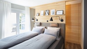 Quarto moderno com cabeceira de madeira, uma janela grande com cortinas leves e prateleira fixada na parede com obras de arte emolduradas.