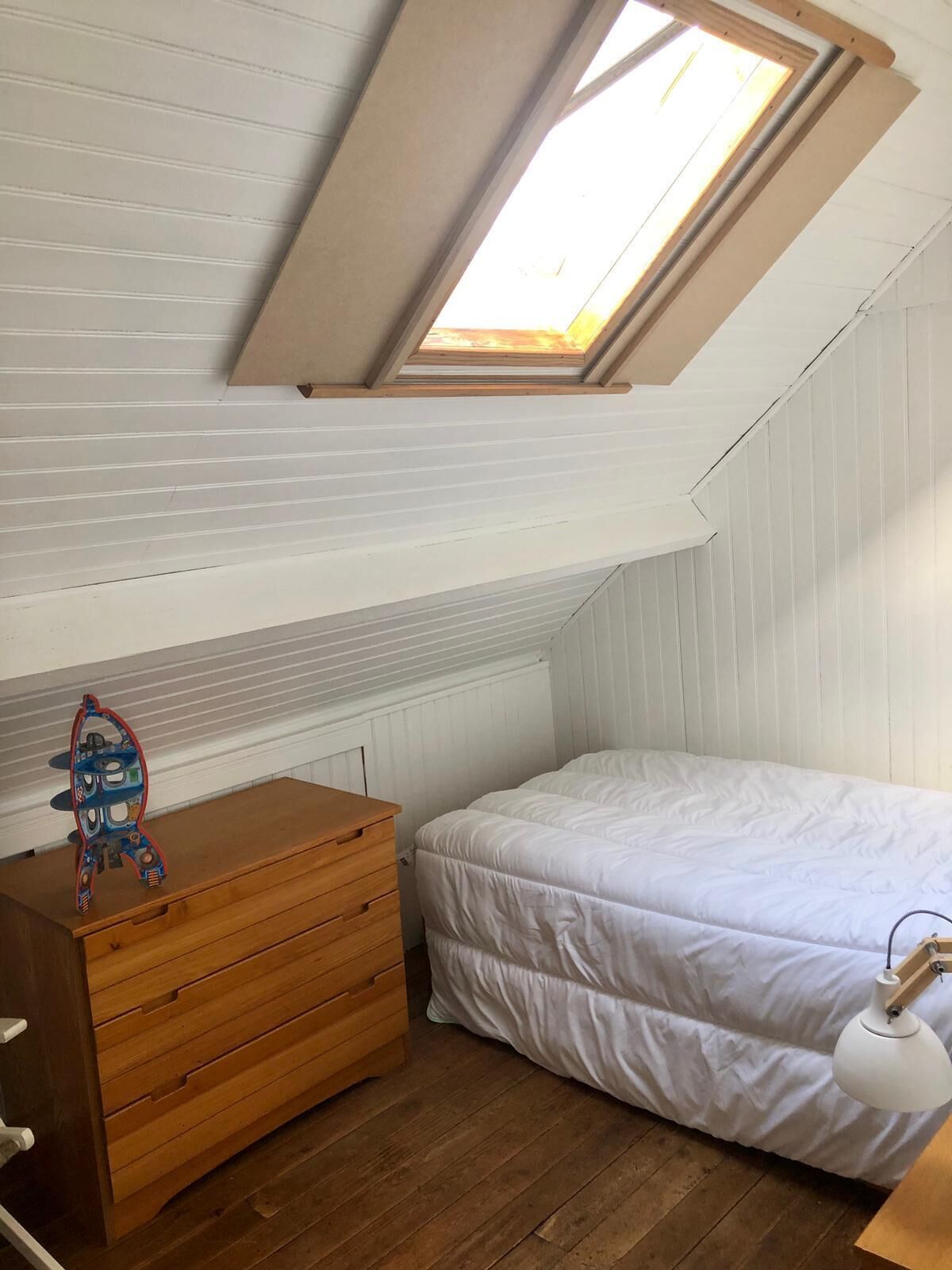 Quarto com claraboia, cómoda de madeira, cama com roupa de cama branca e abajur.