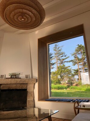 A modern living room with a fireplace, a large window with a view of trees, and a woven ceiling light fixture.