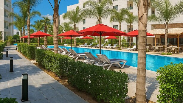 A pool area with lounge chairs and red umbrellas, surrounded by palm trees and a modern building.