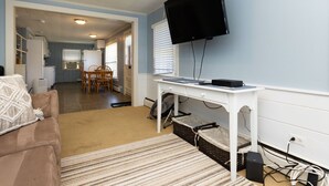 A living room with a flat-screen TV mounted on the wall, a white desk, a brown couch, and a striped rug. - Quintessential Cape Cottage. Block from the beach (Falmouth)