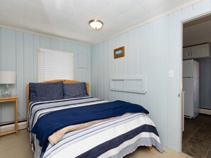 A bedroom with a bed, a nightstand, a window with blinds, a door leading to a kitchen, and a small picture on the wall. - Quintessential Cape Cottage. Block from the beach (Falmouth)