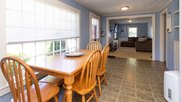 A dining area with a wooden table and chairs, a window with blinds, and a kitchen area in the background. - Quintessential Cape Cottage. Block from the beach (Falmouth)