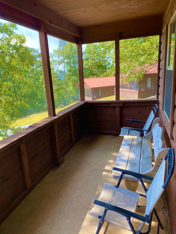 A wooden cabin with a covered porch, large windows, and a view of a red-roofed cabin in the distance.