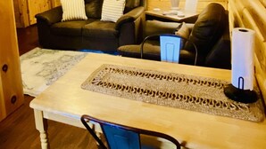 A cozy cabin interior with a wooden wall, a ceiling fan, a framed animal picture, a dark leather sofa, a small table with a lamp, and a blue chair.