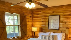 A wooden cabin bedroom with a bed, a nightstand, a lamp, a ceiling fan, a window, and a wall with a framed message.