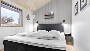 Een net opgemaakt bed met witte lakens, een zwart hoofdbord en een nachtkastje met een lamp. Er is een raam met uitzicht op bomen, een wandplank met boeken en een dromenvanger, en ingelijste foto's aan de muur.