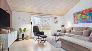 Een moderne woonkamer met een houten plafond, een zwarte bureaustoel, een beige bank en een groot raam met uitzicht op bomen.