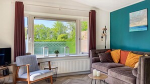A living room with a sofa, a chair, a TV, a window with curtains, a painting, and a radiator. - Gorgeous home in Heinkenszand with WiFi (Heinkenszand)