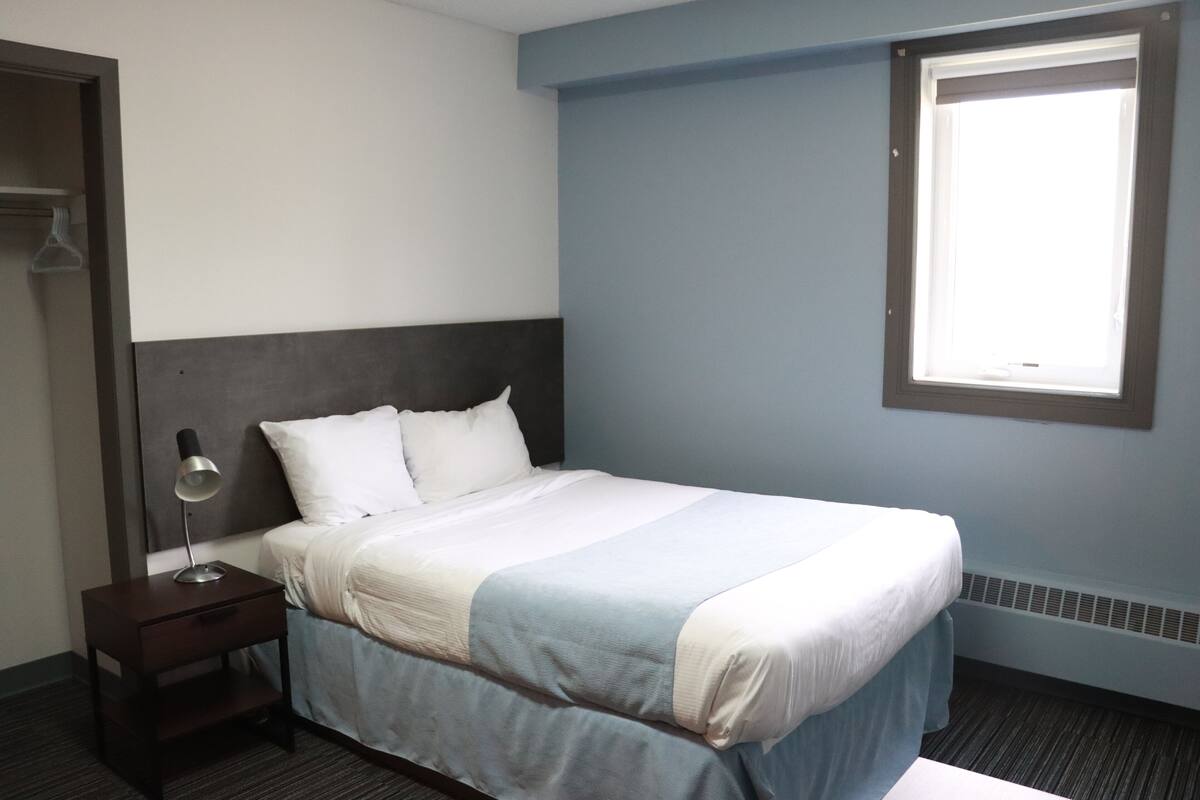 A hotel room with a bed, a nightstand, a lamp, a window, and a wall-mounted heater.