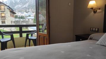 Habitación de hotel con una cama, una mesita, dos sillas, una mesita de noche y una lámpara de pared. Un ventanal grande ofrece vistas a una montaña y un pueblo.