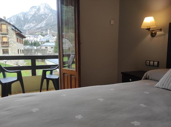 A hotel room with a bed, a small table, two chairs, a nightstand, and a wall-mounted lamp. A large window offers a view of a mountain and a village. - Snö Bielsa (Bielsa)