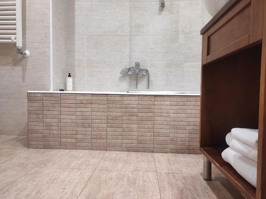 Un baño con pared de ducha azulejada, un lavamanos y un estante de madera con toallas.