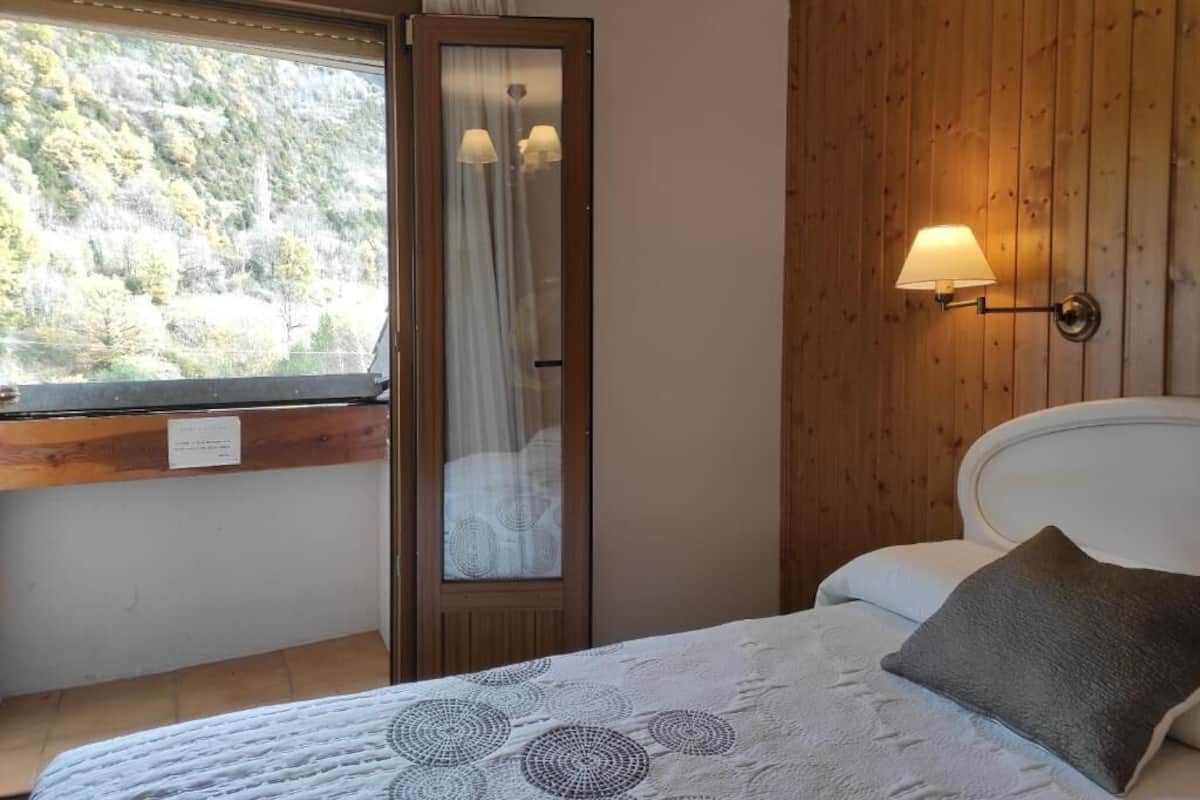 A bedroom with a bed, a window with curtains, a wooden wall, and a wall-mounted lamp.