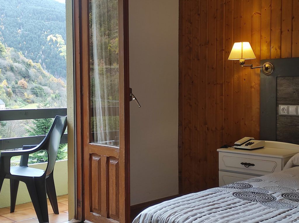 Un dormitorio con cama, una mesita de noche con lámpara, una silla y vistas a un área boscosa a través de un ventanal.