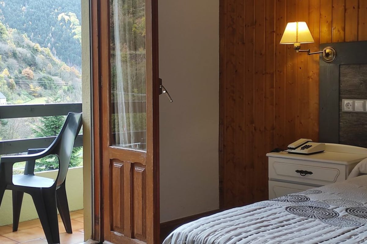 A bedroom with a bed, a bedside table with a lamp, a chair, and a view of a forested area through a glass door.