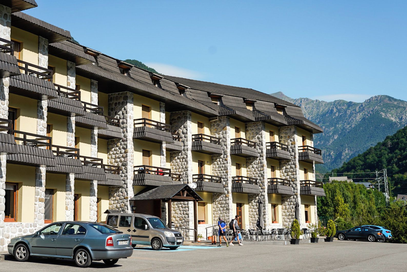 Un hotel de varios pisos con pilares de piedra y balcones, un estacionamiento y montañas al fondo.