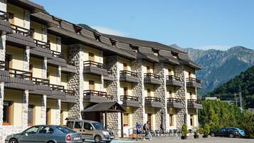 Un hotel de varios pisos con pilares de piedra y balcones, un estacionamiento y montañas al fondo.