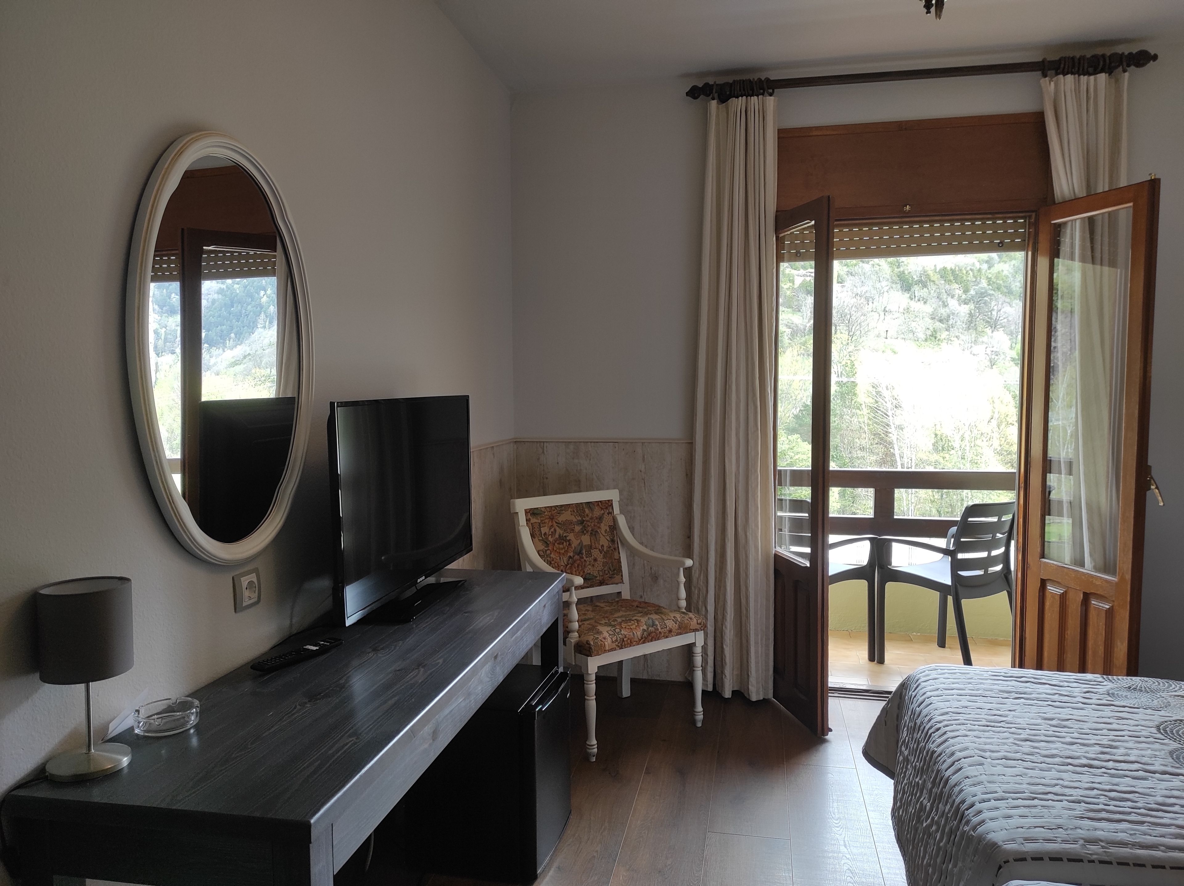 A hotel room with a bed, a TV on a wooden desk, a chair, a balcony with a table and chairs, and a large mirror on the wall.
