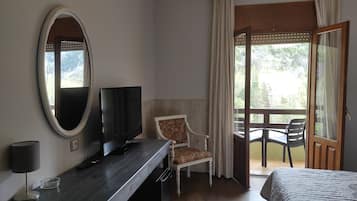 A hotel room with a bed, a TV on a wooden desk, a chair, a balcony with a table and chairs, and a large mirror on the wall.