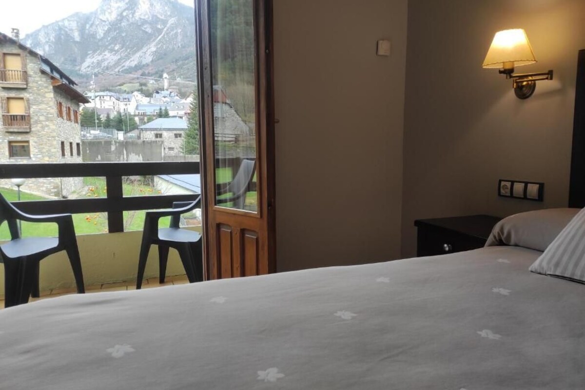 A hotel room with a bed, a small table, two chairs, a nightstand, and a wall-mounted lamp. A large window offers a view of a mountain and a village.