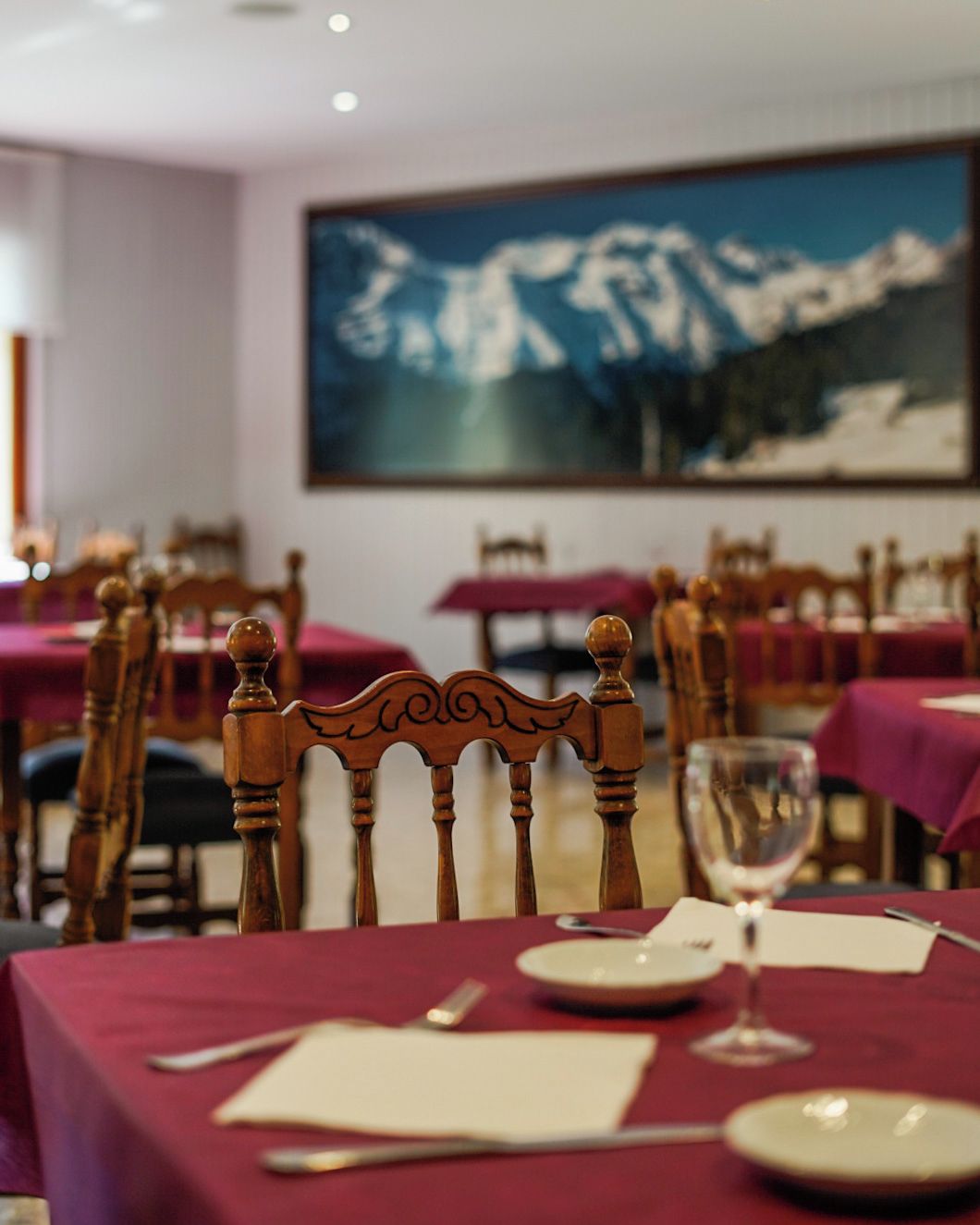 Un comedor con mesas dispuestas para dos, sillas de madera y un cuadro grande de un paisaje montañoso en la pared.