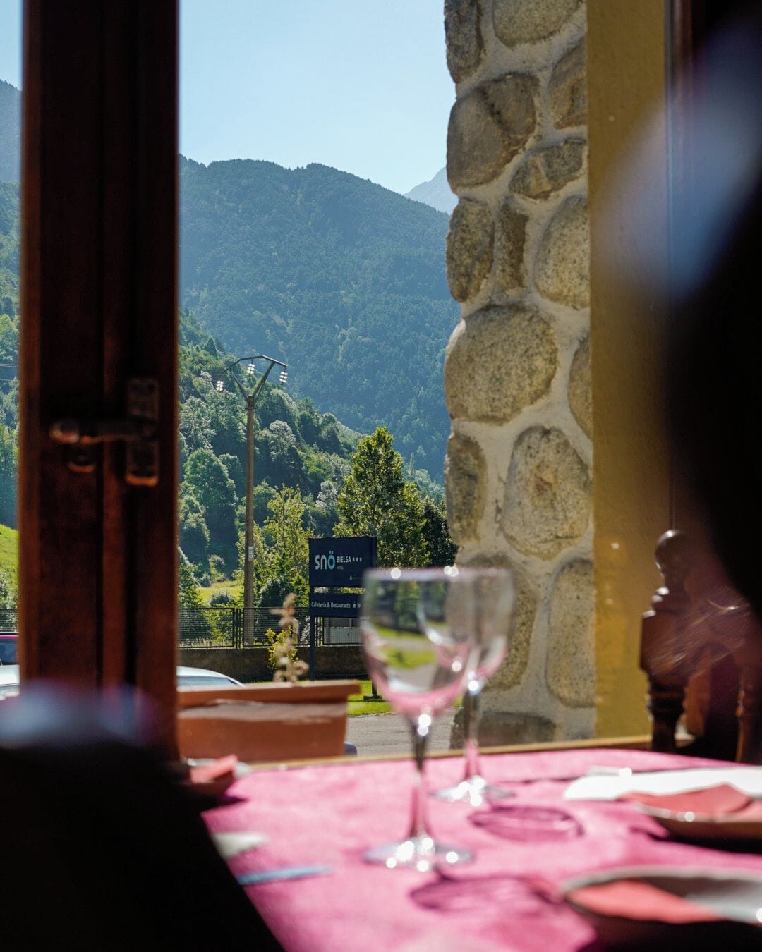 A table with a wine glass, a stone wall, and a view of a mountain landscape.