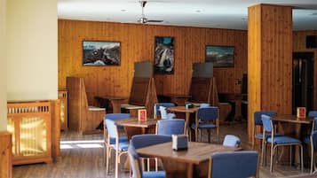 A restaurant with wooden panel walls, blue chairs, and tables. There are paintings on the walls and a ceiling fan.